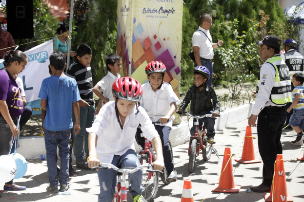 Kinder fahren Fahrräder auf einer Straße mit Hütchen, einige tragen Helme, andere stehen daneben, mit einem Banner, Bäumen und Gebäuden im Hintergrund.