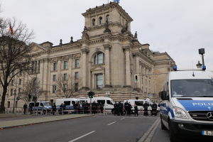 Eine Gruppe von Polizisten steht vor dem Reichstaggebäude in Berlin, Deutschland, mit Fahrzeugen, einem Zaun, Verkehrszeichen, Laternenpfählen, Bäumen und Flaggen im Bild, unter einem klaren Himmel.