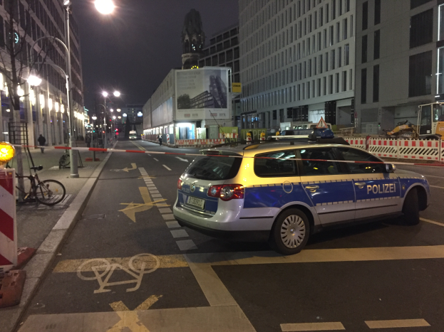 Ein Polizeiwagen steht nachts an einer Straße in Berlin, Deutschland, umgeben von Gebäuden, Straßenlaternen, Verkehrsschildern, Fahrzeugen, Absperrgittern, Fahrrädern, Menschen auf dem Gehweg, Werbetafeln und einer Statue im Hintergrund, wahrscheinlich nach einer Schießerei und einem Autounfall.