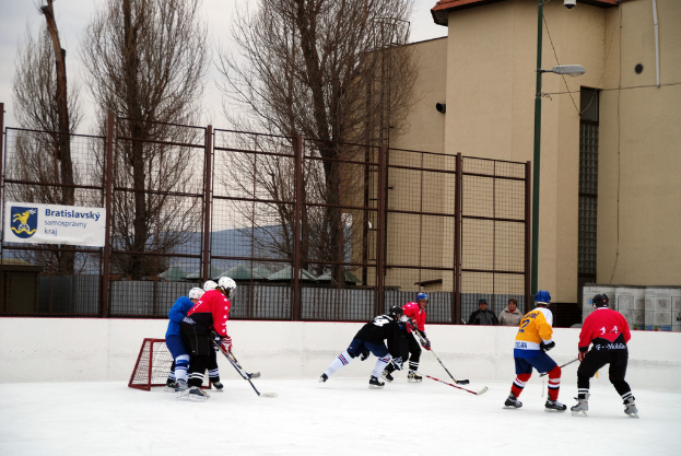 Menschen spielen Eis-hockey auf einer Eisbahn mit Geb√§uden, B√§umen, einer Stra√čenlaterne, einem Namensschild und Z√§unen im Hintergrund unter einem Himmel.