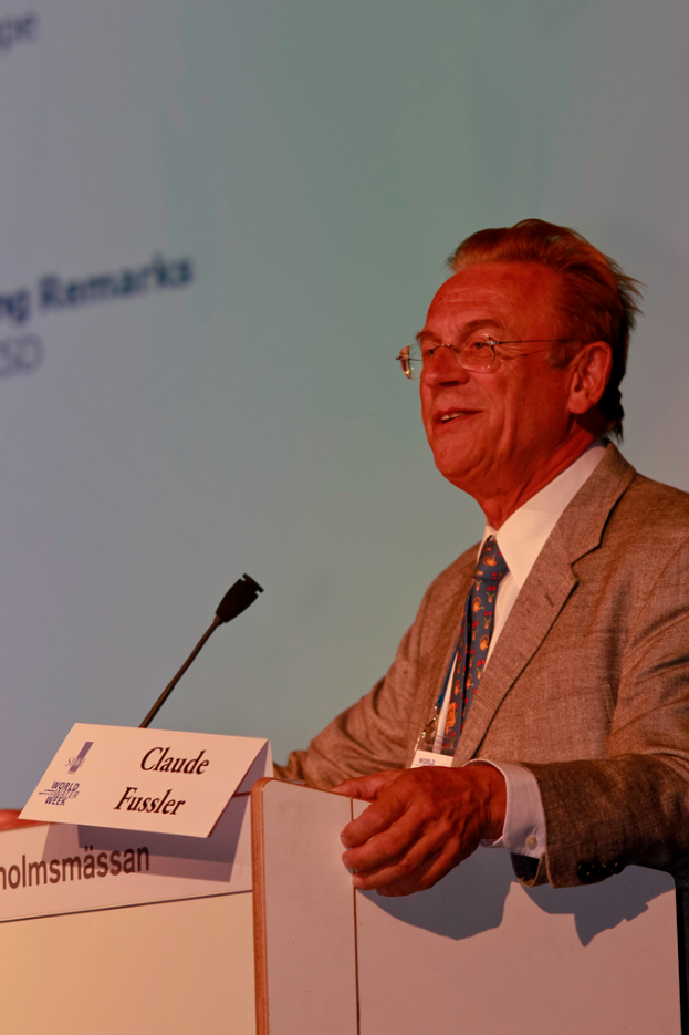Ein Mann in einem weißen Hemd und einem grauen Blazer steht lächelnd da, trägt eine Brille und steht vor einem Podium mit Mikrofon und Namensschildern, mit einem Projektionsschirm oder einer Banner im Hintergrund.