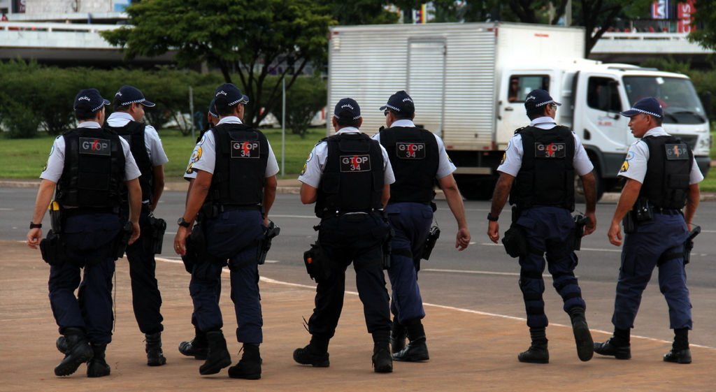 Eine Gruppe von Polizisten in Uniform, einschließlich Mützen, Hemden, Schutzwesten, Uhren, Hosen und Schuhen, die auf einer Straße mit einem geparkten Lkw auf der rechten Seite neben einem Zaun, Gebäuden, Bäumen, Pflanzen, Pfählen und Bannern im Hintergrund und einer Brücke oben rechts gehen.