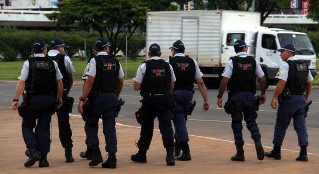 Eine Gruppe von Polizisten in Uniform, einschließlich Mützen, Hemden, Schutzwesten, Uhren, Hosen und Schuhen, die auf einer Straße mit einem geparkten Lkw auf der rechten Seite neben einem Zaun, Gebäuden, Bäumen, Pflanzen, Pfählen und Bannern im Hintergrund und einer Brücke oben rechts gehen.
