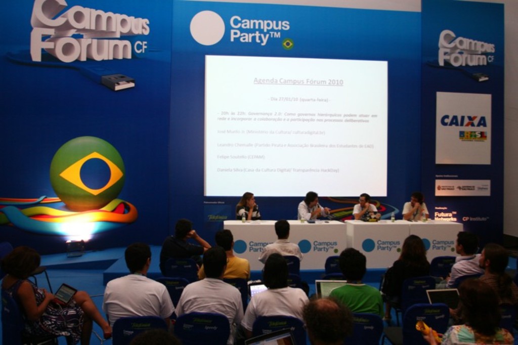 Ein Konferenzsaal mit an Tischen sitzenden Teilnehmern mit Laptops und Wassergläsern, ein Projektionsschirm mit Inhalt und eine blaue Tafel mit Text auf der rechten Seite.