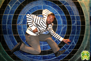 Ein Mann tanzt vor einem bunten Hintergrund mit einem Logo, das Text in der oberen rechten Ecke enthält.