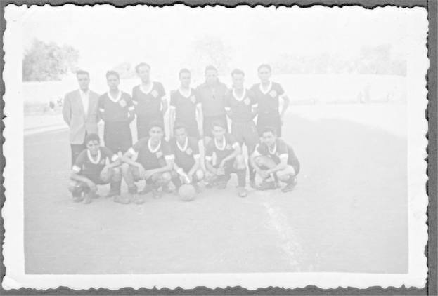 Ein Schwarz-Weiß-Foto in einem Rahmen, das fünf Männer in einer Mannschaftsposition mit einem Ball vor ihnen zeigt, sieben Männer stehen dahinter, Sand am Boden, Bäume und eine Wand im Hintergrund und den Himmel oben.