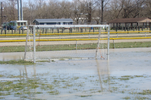 Ein Fußballtor steht auf einem überfluteten Feld mit Gras und Wasser, umgeben von Hütten, Pfosten, Bäumen, Fahrzeugen und einem klaren blauen Himmel im Hintergrund.