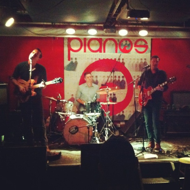 Drei Männer auf der Bühne: Einer spielt Schlagzeug in der Mitte, zwei spielen Gitarre und singen links und rechts daneben, mit einem Banner an der Wand und Lampen an der Decke.