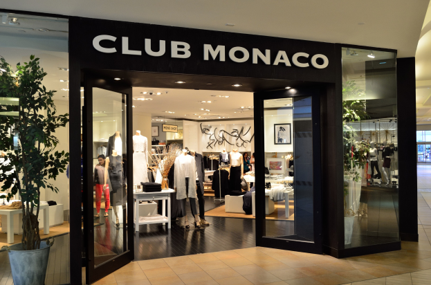 Ein Club Monaco Geschäft in einem Einkaufszentrum mit Glaswänden und -türen, das Schaufensterpuppen in Kleidern, Tische mit Gegenständen, eine Topfpflanze und Deckenleuchten zeigt.