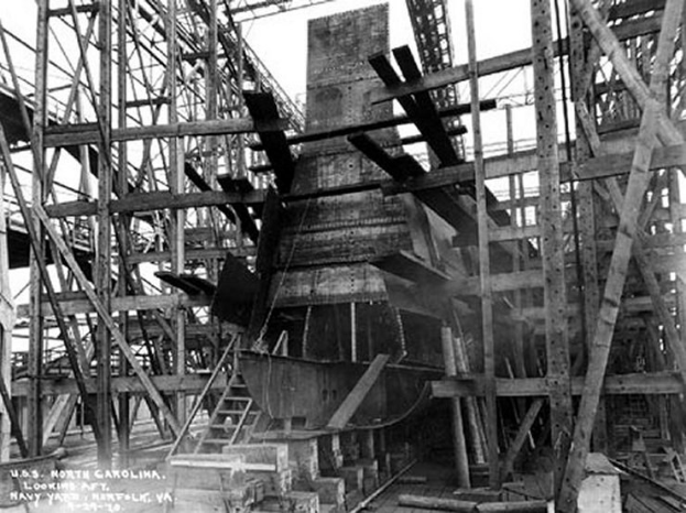 Schwarzes und weißes Foto eines Schiffes in einem Schiffbau, mit hölzernen Pfählen und Treppen im Vordergrund und Himmel im Hintergrund; Text "Marinebewaffnung" in der linken unteren Ecke.