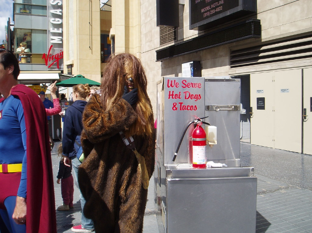 Menschenmenge vor Gebäude mit sichtbarer Beschriftung und Türen, mit einer Box und einem Feuerlöscher in der Nähe.