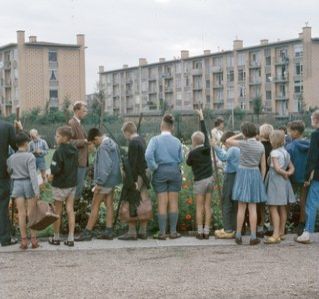 Eine Gruppe von Kindern vor einem Gebäude mit blühenden Pflanzen und Bäumen unter einem klaren blauen Himmel, einige tragen Schulranzen.