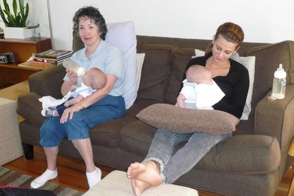 Zwei Frauen sitzen auf einem Sofa und halten Babys, mit Kissen dazwischen, vor einer weißen Wand, mit einem Schreibtisch, Büchern und einer Zimmerpflanze links daneben.