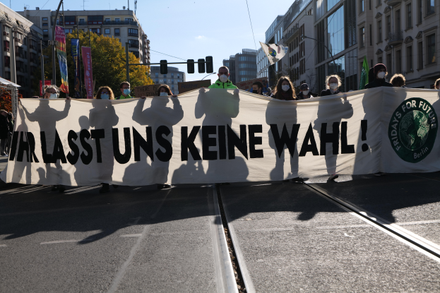 Eine Gruppe von Menschen mit Masken hält ein Banner mit der Aufschrift "Wir sind die letzten keine wahl" vor Gebäuden, Bäumen, Laternen, Ampeln und einem Zelt, mit dem Himmel im Hintergrund.