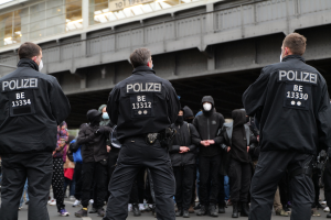 Polizeibeamte in schwarzen Uniformen und Masken stehen vor einer Menge wöhrend einer Demonstration, mit einer Brücke und einem Gebäude im Hintergrund.