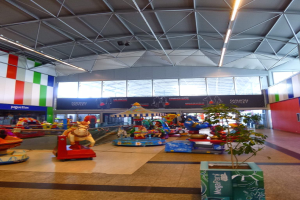 Großer Indoor-Spielplatz in einem Einkaufszentrum mit verschiedenen Fahrgeschäften, einem Topfpflanze, verstreuten Spielzeugen, Informationsschildern, Deckenleuchten und Metallstützpfosten.