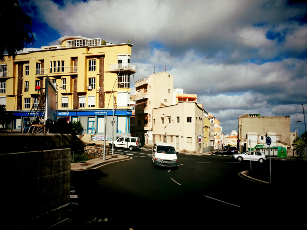 Eine Stadtstraßenszene mit Fahrzeugen, Pfählen, Pflanzen, Schildern und Straßenlaternen im Vordergrund, Gebäuden, Autos, Türen, Fenstern und einem Baum in der Mitte, unter einem teilweise bewölkten, sonnigen Himmel.