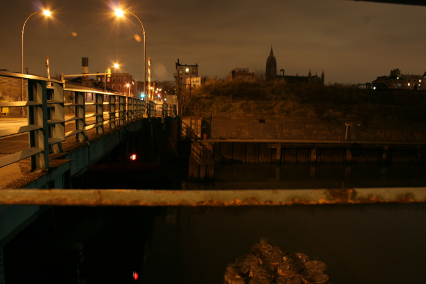 Eine Brücke spannt einen Fluss mit mehreren Straßenlaternen darauf und zahlreiche Gebäude sind im Hintergrund zu sehen.