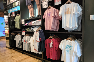 Eine Disney-Store-Ausstellung voller verschiedener T-Shirts an Bügeln und Tafeln mit Text und Bildern von Menschen, mit einem Bildschirm auf der linken Seite und dem Boden unten sichtbar.
