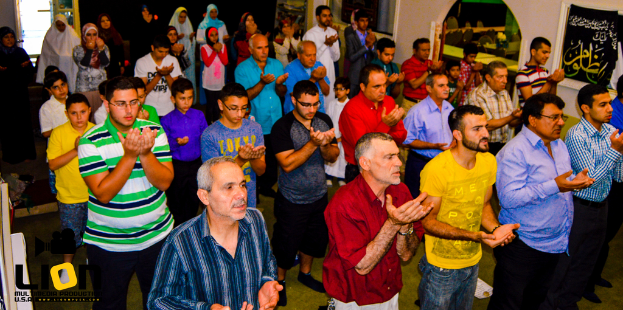 Menschen auf einer Straße mit einem Kreuz und Blumensträußen in der Mitte, fahrende Autos dahinter und Gebäude, Bäume und Straßenlaternen im Hintergrund.