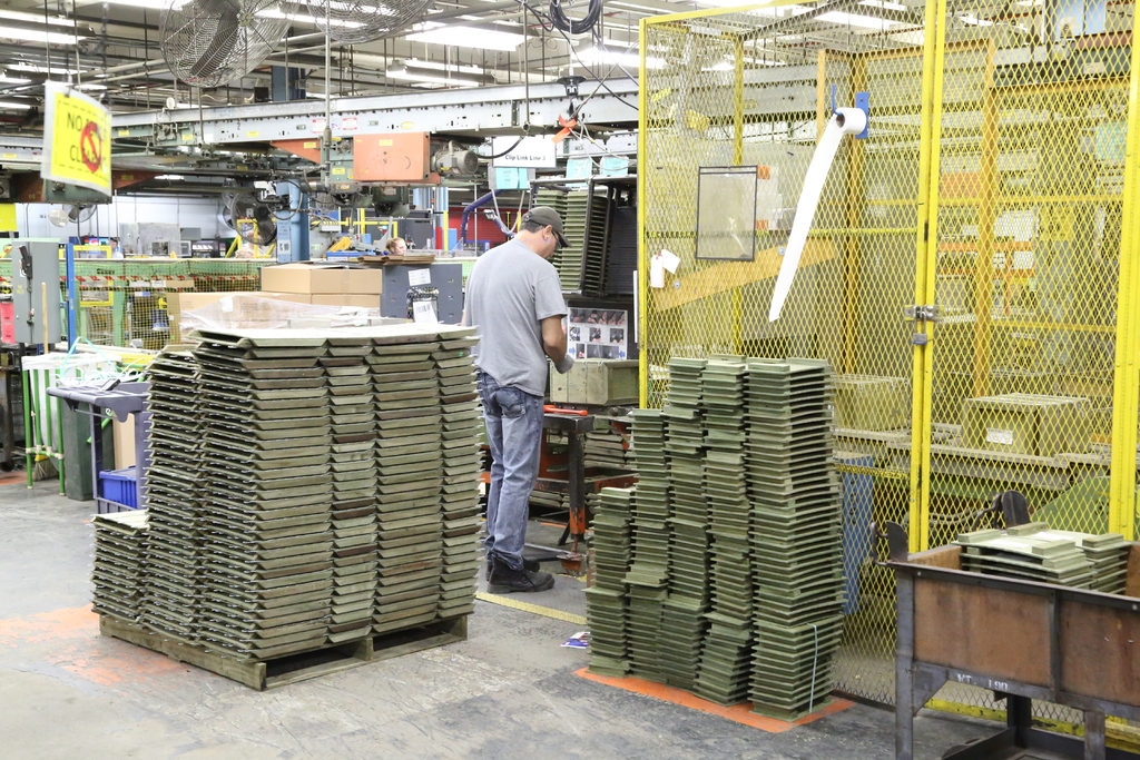 Ein Mann in einer Mütze und T-Shirt steht in einer Fabrik, arbeitet in der Nähe von Maschinen auf dem Boden und an der Decke, mit Lüftern und Lampen ebenfalls sichtbar.