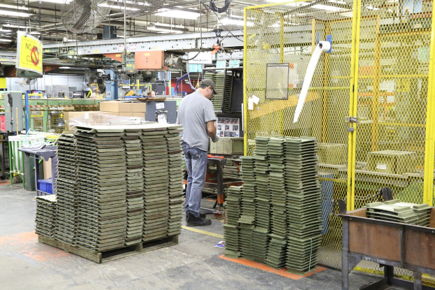 Ein Mann in einer Mütze und T-Shirt steht in einer Fabrik, arbeitet in der Nähe von Maschinen auf dem Boden und an der Decke, mit Lüftern und Lampen ebenfalls sichtbar.
