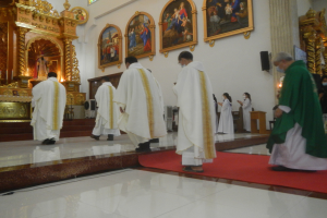 Eine Gruppe von Priestern geht einen roten Teppich in einer Kirche entlang, mit Menschen auf dem Boden und auf der rechten Seite, und Fotorahmen an der Wand im Hintergrund.