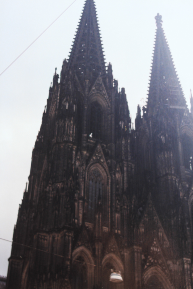 Der Kölner Dom in Köln, Deutschland, mit seinen Spitzen, die in den Himmel ragen, und ein paar Menschen vor dem Gebäude mit sichtbaren Kabeln im Hintergrund.