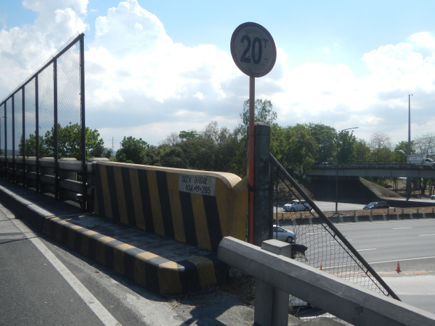 Eine Brücke mit einem Tempolimit-Schild an der Seite, umgeben von Fahrzeugen, einem Zaun, Pfählen, Lampen, Bäumen und einem bewölkten Himmel im Hintergrund.
