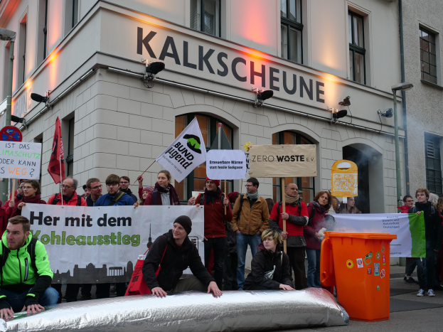 Eine Gruppe von Menschen mit Schildern und Plakaten steht vor einem Gebäude, mit zwei Personen im Vordergrund und einem Müllcontainer auf der rechten Seite, während einer Demonstration in Deutschland, mit Gebäuden, Fenstern, Lichtern und Schildern im Hintergrund.