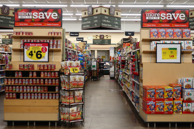 Ein Kiosk-Interieur Mit Regalen Und Boden, Bestückt Mit Verschiedenen Lebensmitteln, Überkopfbeleuchtung Und Informationsschilder Im Hintergrund, Mit Einer Person Im Entfernten.