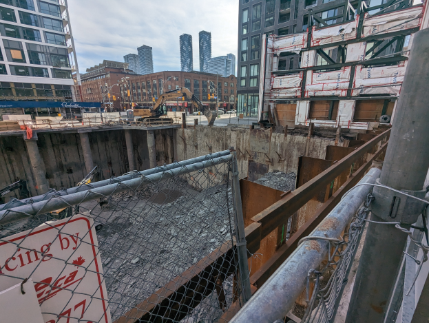 Eine Baustelle mit einem Zaun und einem Schild mit der Aufschrift "Racing by CCA", umgeben von Gebäuden, Fahrzeugen auf der Straße und einem bewölkten Himmel.