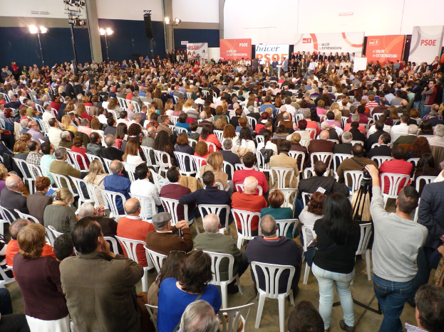 Ein volles Stadion mit vielen sitzenden und stehenden Menschen, eine Buhne mit zwei Rednern am Podium und mehrere Plakate und Lichter in der Nahe.