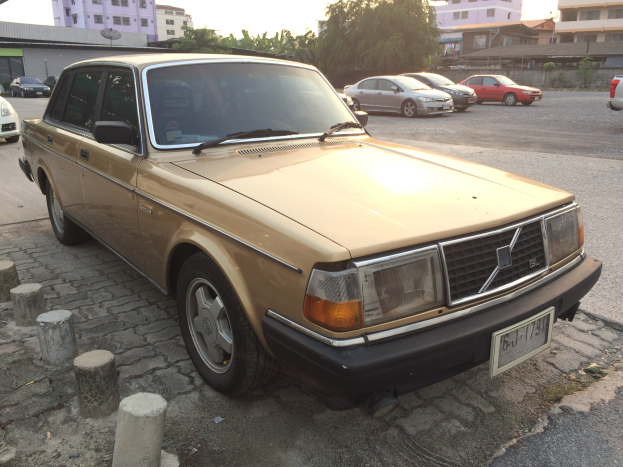 Ein goldfarbener Wagen am Straßenrand mit Gebäuden, Bäumen und einem klaren blauen Himmel im Hintergrund.