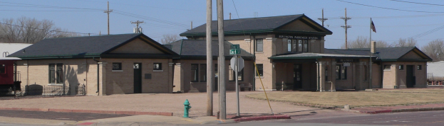 Ein Bahnhofsgebäude mit einem Feuerhydranten davor, umgeben von Häusern, einem Zaun, Bäumen, Strommasten mit Drähten, Schildern, einer Flagge an einer Fahnenstange, unter einem bewölkten Himmel.