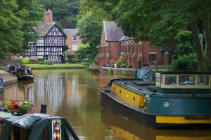 Kanal mit mehreren ankernden Booten, umgeben von grünem Gras, Pflanzen und Bäumen, mit Gebäuden im Hintergrund.