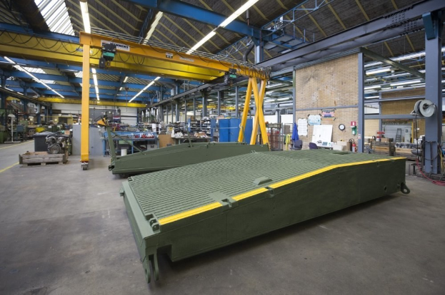 Großes Lager mit grünen Behältern, Maschinen, Polen, Beleuchtung und Stangen; Hintergrund zeigt eine Wand mit einer Uhr und Papieren.