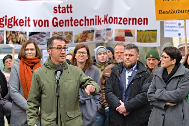 Ein Mann spricht in ein Mikrofon auf einer öffentlichen Veranstaltung, umgeben von einer Menge, mit einem Banner und Transparent-haltenden Personen dahinter.
