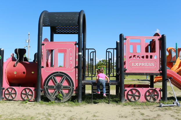 Ein Kind klettert Treppen an Spielgeräten mit Bäumen, Polen, Stromleitungen und Wolken im Hintergrund und einem Roller auf dem Boden rechts.