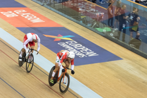 Zwei Männer mit Helmen rasen auf Rennrädern auf einer Indoor-Bahn vor Publikum, mit einer Glasscheibe, auf der 'Europameisterschaften Glasgow 2018' steht und Zuschauern dahinter.