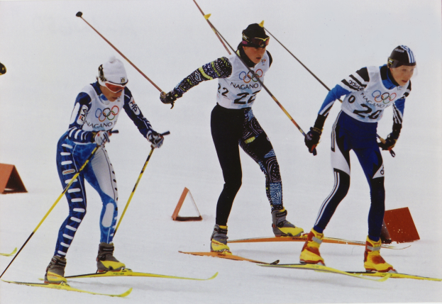 Drei Skifahrer sausen eine schneebedeckte Piste hinunter, jeder hält Ski-Stöcke in der Hand, trägt bunte Ski-Anzüge und Helme, während die Sonne auf dem Schnee glitzert.
