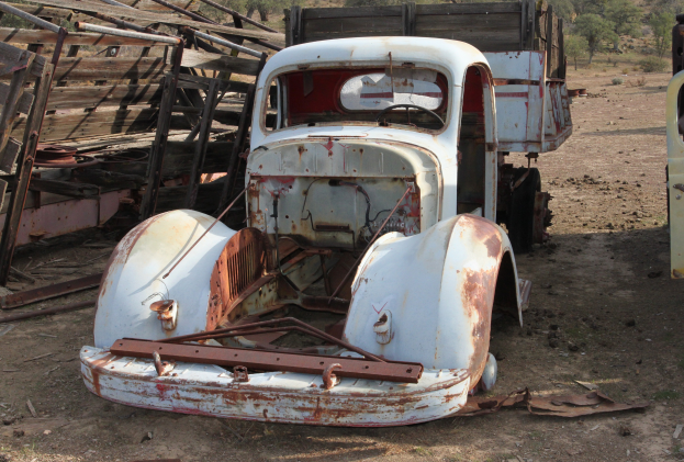 Ein alter, rostiger Lastwagen in einem schlechten Zustand steht im Schmutz, umgeben von einem Holzzaun und Bäumen im Hintergrund.