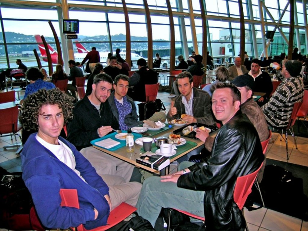 In einem Flughafen sitzen eine Gruppe von Menschen um einen Tisch mit roten Stühlen und essen und unterhalten sich, wobei einer einen blauen Pullover und ein anderer eine schwarze Jacke und Jeans trägt und ein schwarzer Koffer und ein Glas, ein Hügel, ein Flugzeug und ein Fernseher im Hintergrund zu sehen sind.