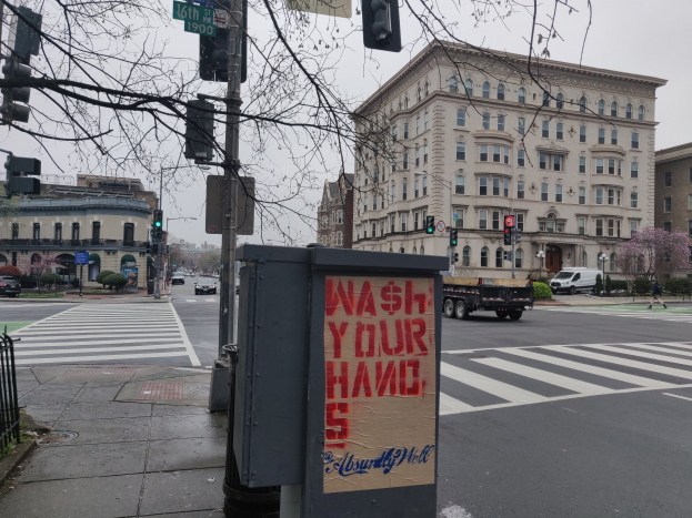 Eine belebte Stadtstraße mit Fahrzeugen, einem Fußgänger, einem Müllcontainer mit einer 'Waschen Sie Ihre Hände'-Aufschrift, Verkehrsampeln, Hinweisschildern, einem Metallzaun, Pflanzen, Bäumen, Gebäuden und einem klaren blauen Himmel.
