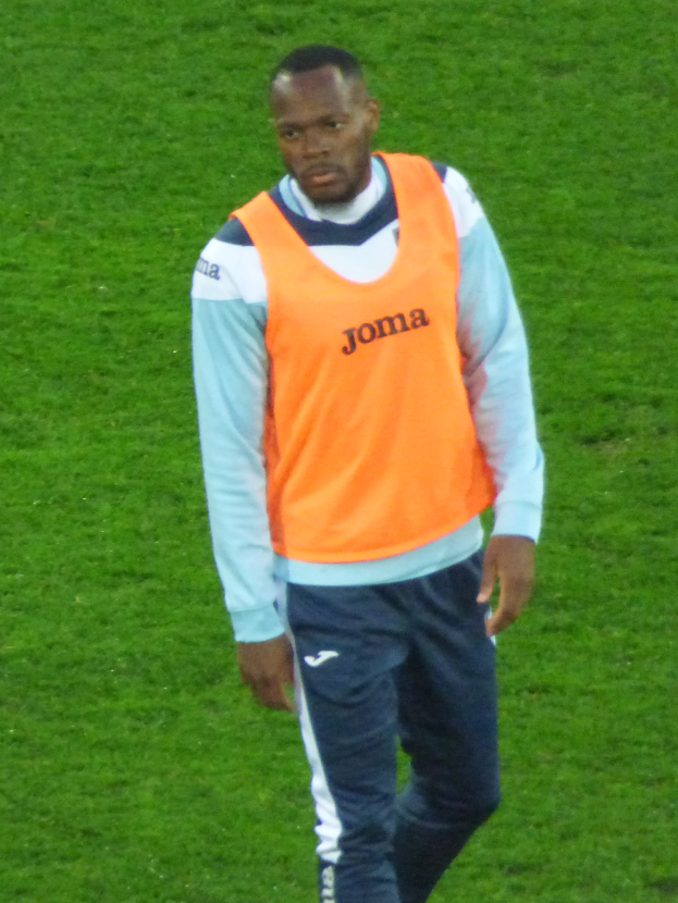 Ein Mann in einem orangen Hemd und blauen Hosen geht entschlossen über ein grünes Feld.