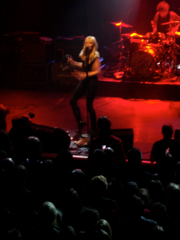 Eine Frau singt auf der Bühne in ein Mikrofon, während ein Mann daneben Schlagzeug spielt, mit Zuschauern im Publikum.