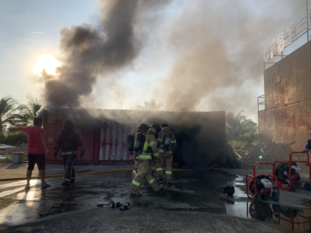 Feuerwehrleute in Helmen arbeiten daran, ein Gebäude Feuer zu löschen, während Rauch aufsteigt, mit einem anderen Gebäude und Bäumen in der Nähe.