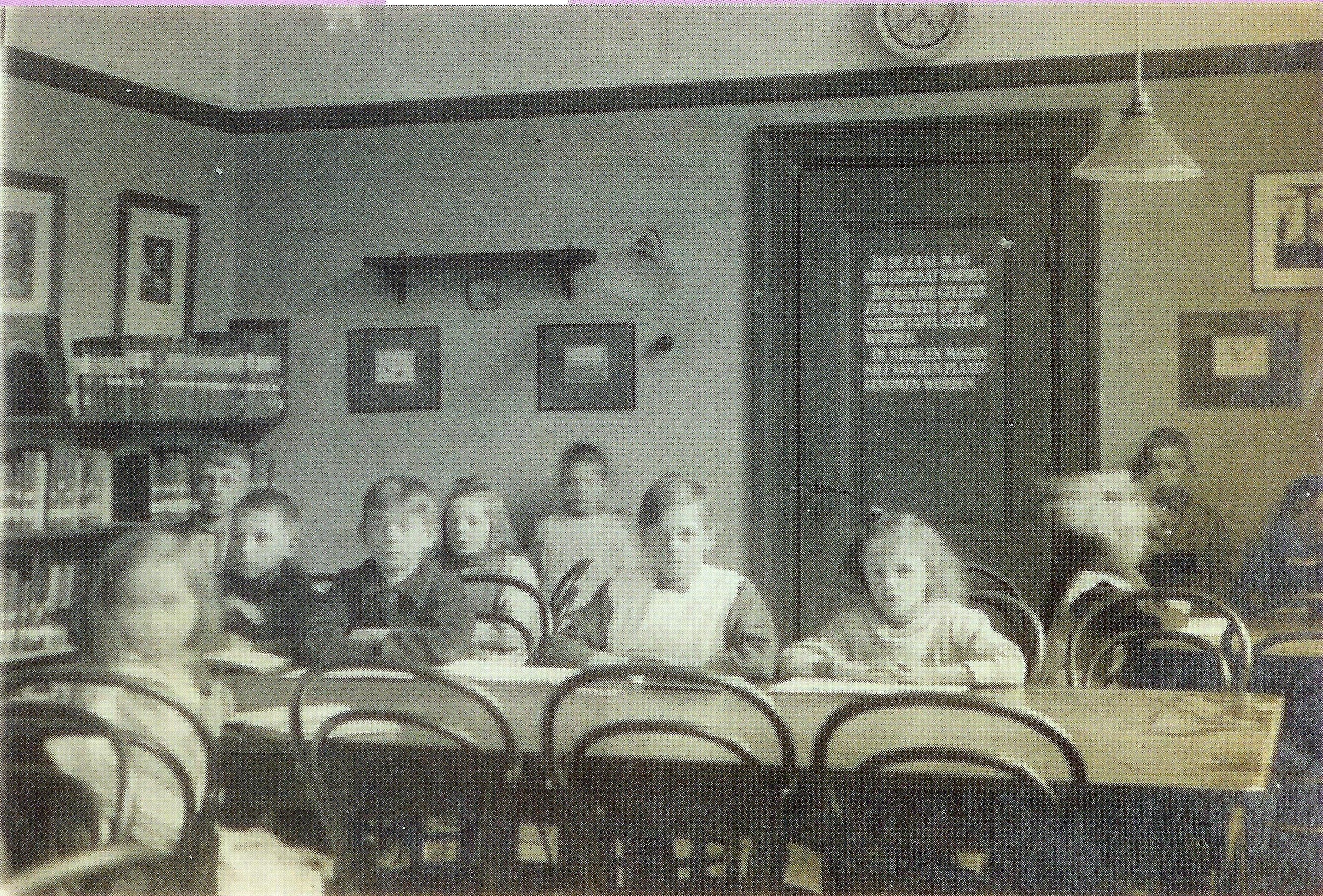 Eine Gruppe von Kindern sitzt an Tischen in einem Klassenzimmer mit Büchern auf den Tischen, Bücherregalen links, Fotorahmen und einer Uhr an der Wand, einer Tür mit Text und Deckenlampen, die für warmes Licht sorgen.