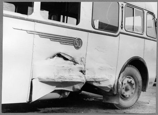 Ein Schwarz-Weiß-Foto eines beschädigten Busses am Straßenrand, dessen Vorderseite eingedrückt ist, Fenster zerbrochen und die Fahrertür öffnet.