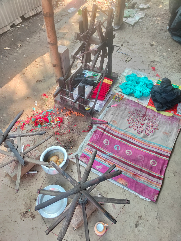 Ein hölzernes Spinnrad auf schmutziger Erde neben einem Haufen Kleidung, Tuch, Schüssel, Teller und anderen Gegenständen, mit einem Baumstamm, einer Wand und zusätzlichen Gegenständen im Hintergrund, was auf eine Textilmühle in Indien hinweist.